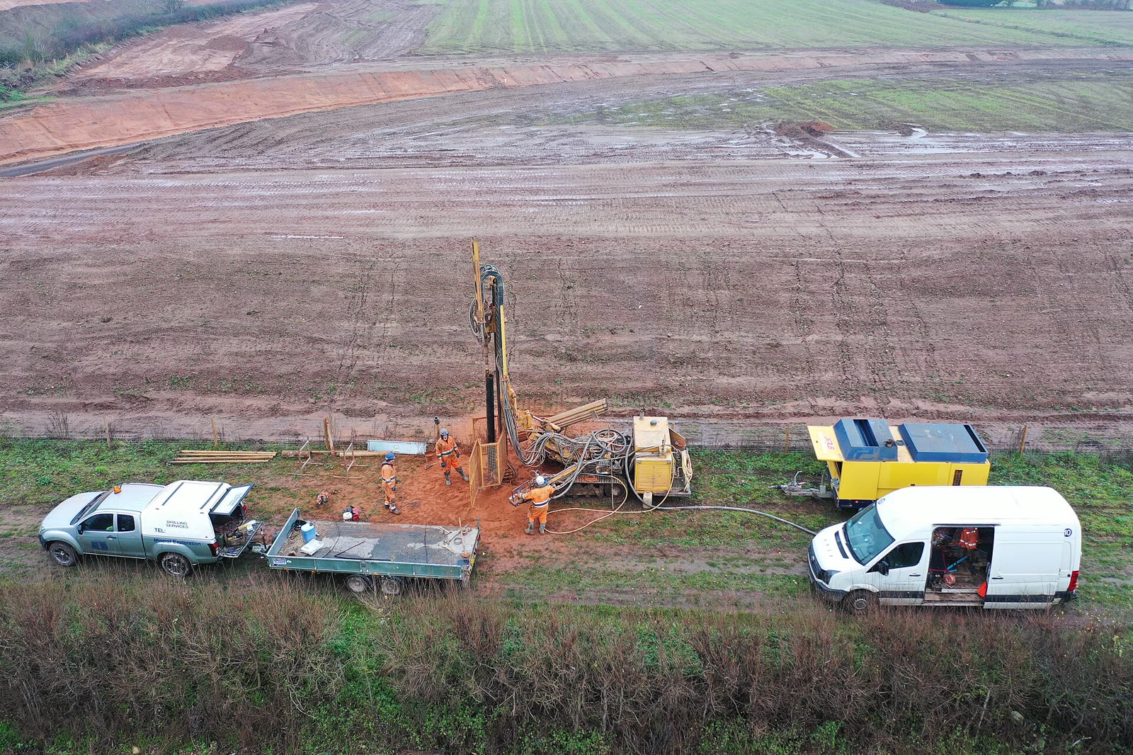 Vista do alto de uma perfuração num campo, com a máquina, um compressor e duas caminhonetes